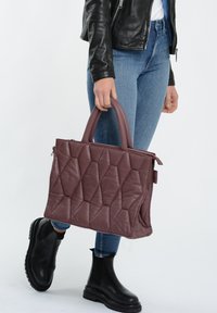 Quiltet bordeaux læderhåndtaske med to runde hanke, lynlås og et geometrisk mønster. Parret med blå jeans og sorte ankelstøvler.