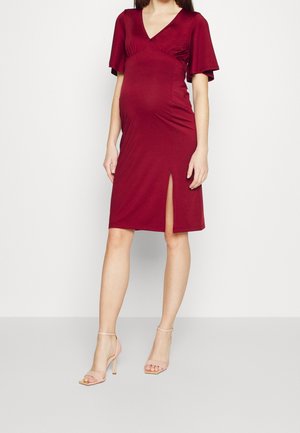 Femme enceinte portant une robe bordeaux mi-longue à manches courtes avec une fente latérale, associée à des sandales à talons hauts couleur nude.