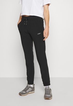 Personne portant un pantalon de jogging noir avec des poignets élastiques et un t-shirt court blanc, debout les mains dans les poches et des baskets grises.