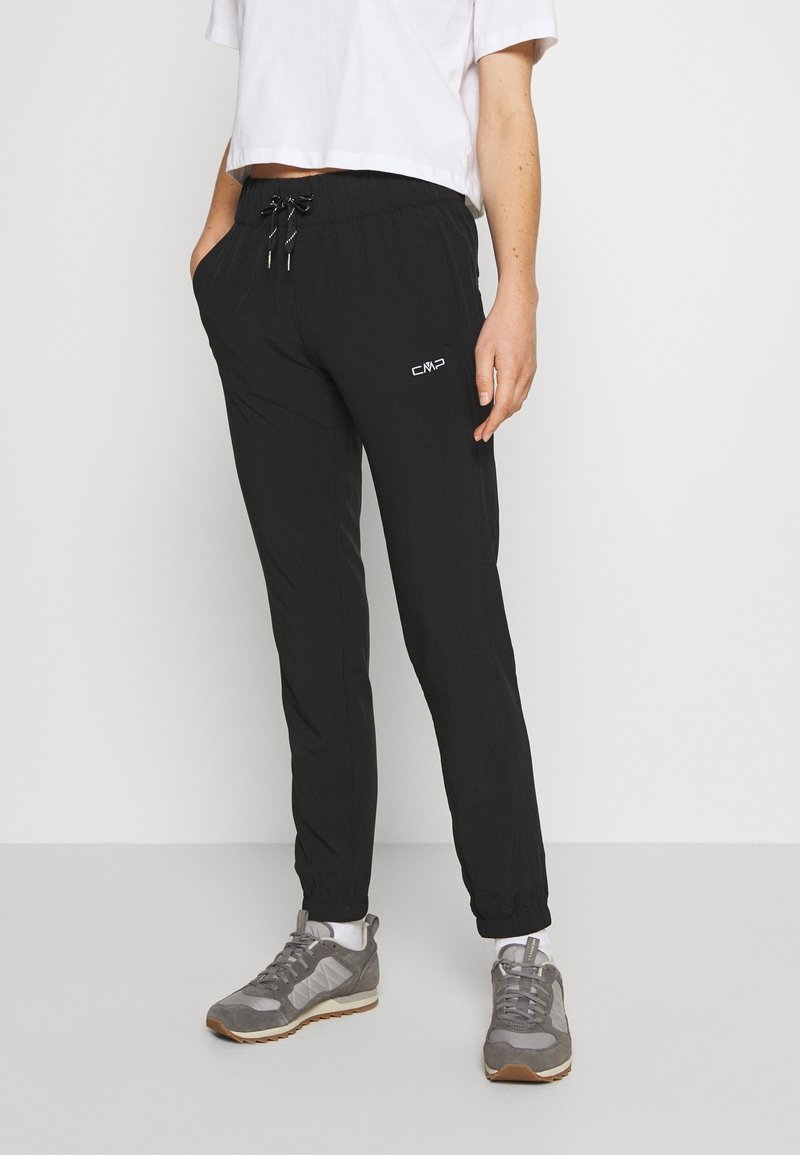 Personne portant un pantalon de jogging noir avec des poignets élastiques et un t-shirt court blanc, debout les mains dans les poches et des baskets grises.