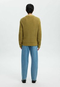 Man som står med ryggen mot kameran och bär en olivgrön grovstickad tröja, lösa ljusblå jeans och svarta skor framför en vit bakgrund.