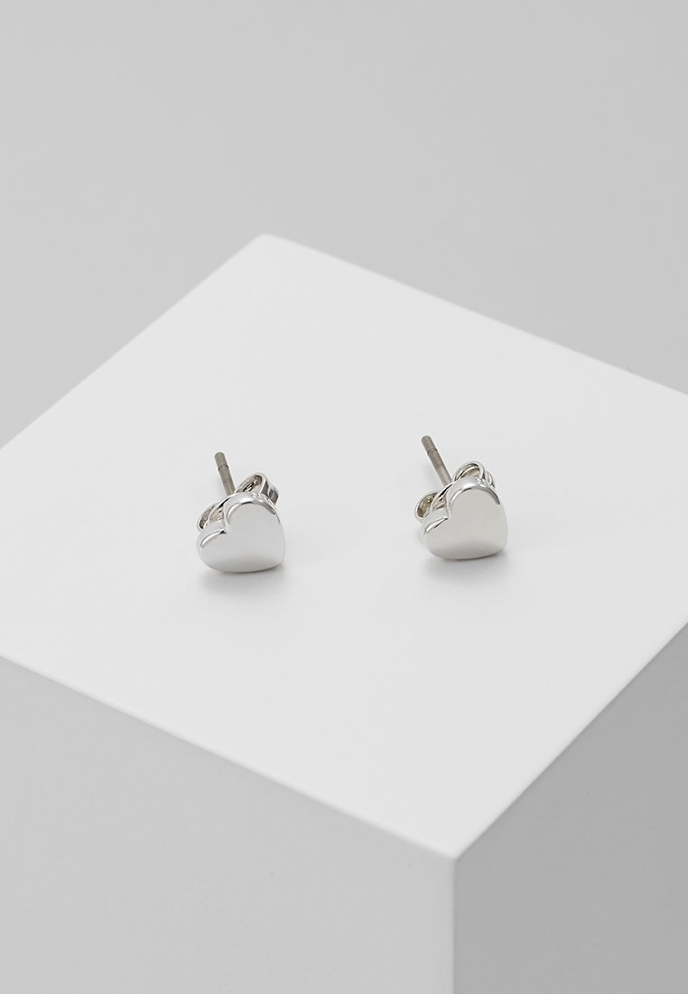 Petites boucles d'oreilles clous en argent en forme de cœur exposées sur un cube blanc sur un fond gris.