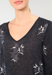 Blusa nera floreale con scollatura a V, caratterizzata da pois bianchi sparsi e un tessuto testurizzato con accenti floreali di colore blu scuro.