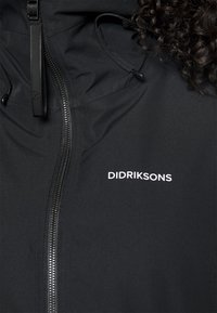 Črna nepremočljiva jakna z zadrgo, nastavljivim kapuco na vrvico in belim logotipom "DIDRIKSONS" na prsih. Gladka tekstura.
