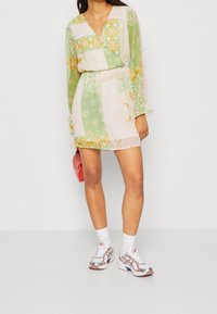 Kobieta w mini sukience w zielono-żółte wzory z długimi rękawami, białe skarpetki, białe i różowe trampki oraz różowa torebka.