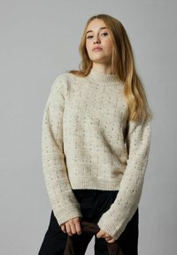 Flauschiger Pullover in Off-White mit strukturiertem Muster, hohem Kragen und gerippten Bündchen. Das Model hält einen braunen Stoff. Der Hintergrund ist in neutralem Grau.