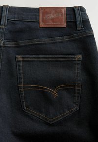 Mørkeblå denim jeans baglomme med orange stikninger og et brunt lædermærke med teksten "Premium Denim Goods" ved taljen.