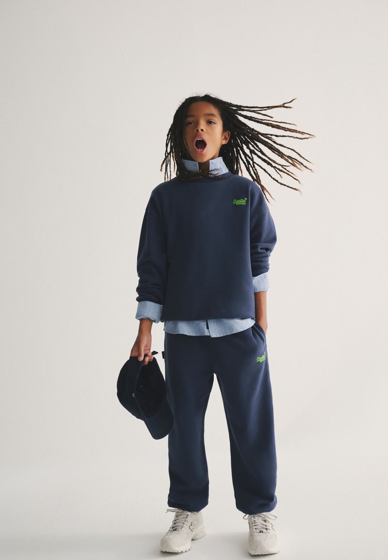 Niño con trenzas largas que lleva sudadera y pantalones azul marino, sosteniendo una gorra en una mano, cabello al viento, de pie frente a un fondo liso.
