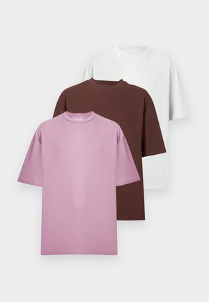 Dos personas con camisetas marrones holgadas y botas negras; una con pantalones negros, la otra con una falda negra, de pie contra una pared blanca.