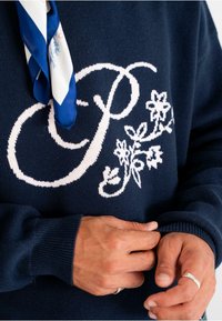 Marineblauer Pullover mit großen weißen gestickten Initialen und floralem Design. Gerippte Bündchen und Kragen. Das Modell justiert den Stoff mit den Händen.