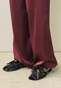 Schwarze Samtslipper mit Blumenmuster, Schleifenakzent, kombiniert mit burgunderroten Satohosen, auf einem strukturierten, hellen Teppich platziert.