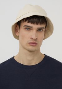 Giovane uomo con pelle chiara, che indossa un cappello a secchiello beige e un maglione girocollo blu navy, che guarda dritto avanti su uno sfondo semplice.