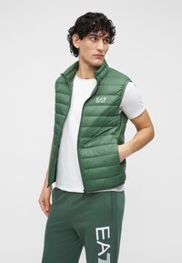 Grön vadderad väst med hög krage och dragkedja, med en liten vit logotyp på vänster bröst. Buren över en vit t-shirt.