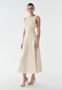 Robe sans manches couleur crème avec une taille smockée, tissu fluide, texture superposée et silhouette jusqu'au genou, associée à des sandales blanches à lanières.