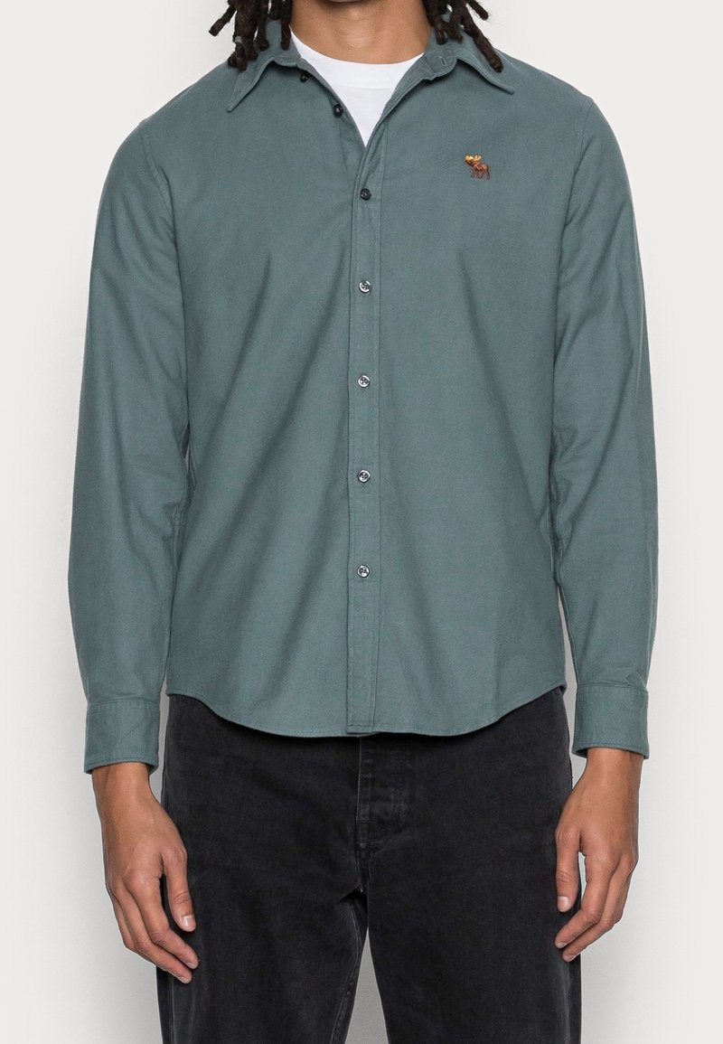 Groen shirt met lange mouwen en een button-down kraag, met een klein bruin geborduurd ontwerp op de borst en knoopsluitingen aan de voorkant.