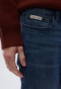 Sötétkék farmernadrág struktúrált felülettel, látható Calvin Klein logóval és rézszínű kovácsolt díszítésekkel. Vörösesbarna pulóverujj is látható.