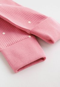 Närbild av rosa ribbstickade ärmar med små vita prickar och enfärgade rosa vikta muddar på vit bakgrund.