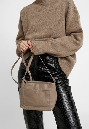 Persona con un suéter marrón de textura peluda y pantalones negros texturizados, sosteniendo un bolso de cuero color topo con bolsillo frontal y correa para el hombro.