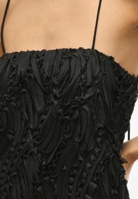 Juoda suknelė, pagaminta iš tekstūruoto juostos tipo audinio su volanų dizainu. Su plonomis petnešėlėmis ir aptemptu liemeniu.