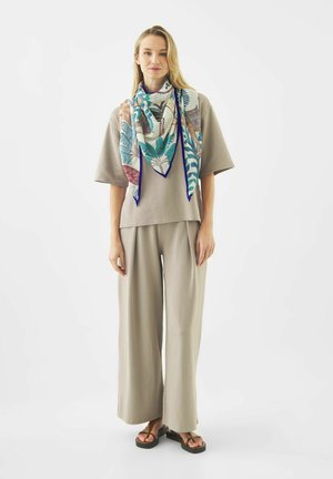 Vrouw draagt een losse beige top en broek, een blauw-witte sjaal met patroon en zwarte sandalen, staand tegen een effen witte achtergrond.