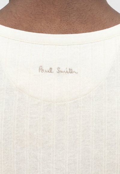 Κοντινή λήψη του πίσω λαιμού ενός ριμπ ύφους τοπ εκρού με το κέντημα "Paul Smith" σε μικρή μπεζ κλωστή.
