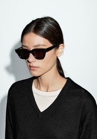 Black sunglasses with a geometric design, dark lenses, and chunky frames. Worn with a black V-neck sweater over a light-colored top.