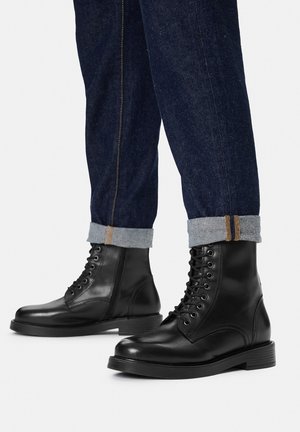 Schwarze Leder-Schnürstiefeletten, kombiniert mit dunkelblauen, aufgekrempelten Jeans, auf weißem Hintergrund.