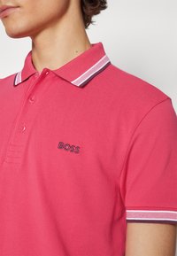 Gros plan d’une personne portant un polo rose vif avec des bordures blanches et foncées au col et aux manches, arborant un petit logo « BOSS » sur la poitrine.