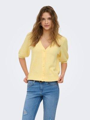 Giovane donna con capelli mossi che indossa un cardigan giallo a maniche corte abbottonato e jeans blu strappati, in piedi con le mani che tirano l'orlo del cardigan.