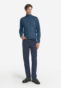 Blauer Rollkragenpullover kombiniert mit dunklen Jeans. Der Pullover hat eine glatte Textur; die Jeans haben einen geraden Schnitt und minimale Nähte.