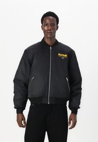 Giubbotto bomber nero con colletto a costine, tasche con zip e un audace logo giallo della Kodak sul petto. Indossato sopra pantaloni neri.