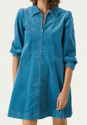 Femme portant une robe en velours côtelé bleu avec une fermeture éclair à l'avant, un col et des manches trois-quarts froncées, devant un fond uni.