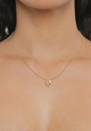 Zarte goldene Halskette mit einem kleinen, gebogenen Anhänger, der mit sieben kleinen klaren Kristallen verziert ist, getragen am Hals einer Person.