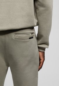 Pantalones jogger de tela suave de color gris claro con un ajuste ceñido, mostrando un bolsillo trasero y una pequeña etiqueta de la marca cerca del bolsillo.