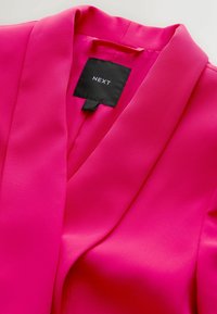 Svetloružové sako s výrezom a čiernou textilnou štítkom s bielym nápisom značky „NEXT“ vo vnútri výstrihu.