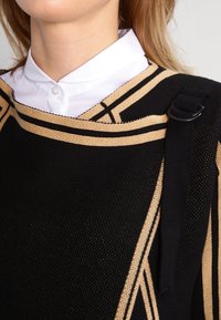 Schwarzes Strickoberteil mit goldenen geometrischen Streifen, rundem Halsausschnitt und einem Schulterträger mit einem Schnallendetail. Weißer Hemdkragen sichtbar.