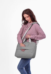 Eine Frau in einem mauvefarbenen Hoodie und Jeans steckt ihre Hand in die vordere Tasche einer grauen Schultertasche mit verstellbarem Trageriemen.