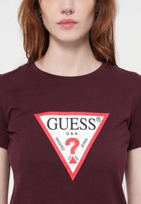 Mujer vistiendo una camiseta burdeos con un gran logo triangular en blanco y rojo de Guess que presenta un signo de interrogación y el texto "Washed Jeans U.S.A."