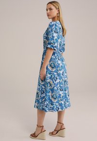 Blå blomstret kjole med puffærmer, knælang, lavet af let stof. Har en indsnøret talje og en let skørt. Kombiner med wedgesandaler.