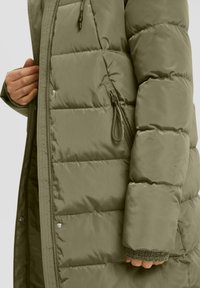 Kadar izbliza osobe koja nosi maslinasto zeleni kaput s podstavom, s vidljivim patentnim zatvaračem i rukom unutar otvorenog prednjeg dijela.