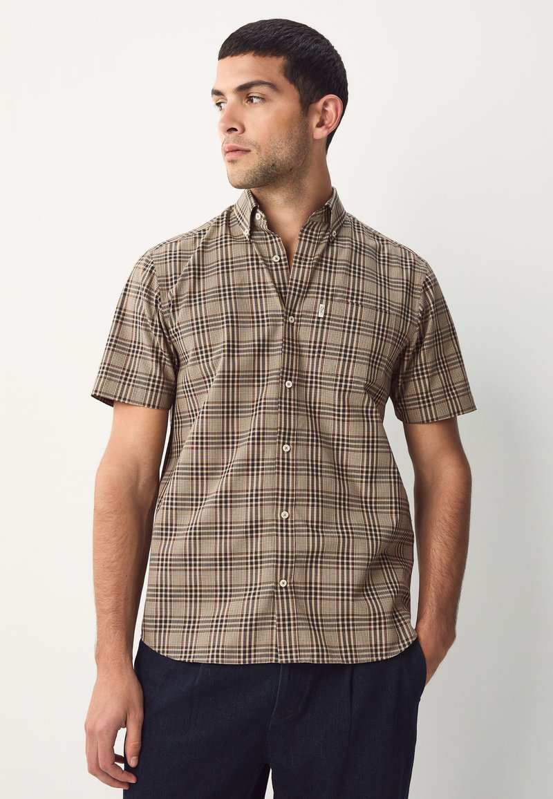 Man with short dark hair wearing a beige and black plaid short-sleeve button-up shirt, standing with one hand in dark pants pocket.