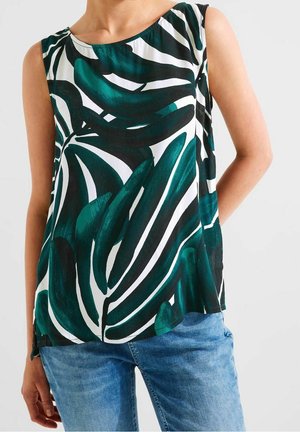 Mouwloze top met grote groene en zwarte abstracte bladpatronen gedragen met blauwe spijkerbroek bij een persoon tegen een effen achtergrond.
