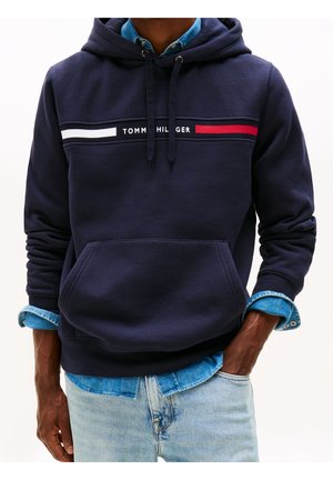 Mężczyzna ubrany w granatową bluzę z kapturem z logo "Tommy Hilfiger" na piersiach, nałożoną na jasnoniebieską koszulę jeansową i jasne jeansy.
