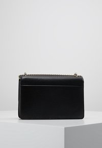 Černá kožená kabelka se stříbrným řetízkovým popruhem, obdélníkového tvaru a plochou kapsou na zadní straně, vystavená na bílém podstavci.