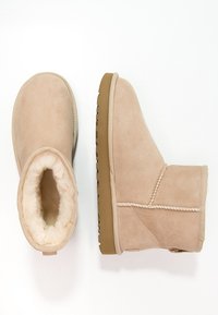Botas de invierno de ante beige con forro de piel sintética suave, una bota mostrando la vista superior y la otra en perfil lateral sobre un fondo blanco.