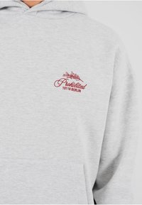 Sudadera gris hecha de una tela suave, con texto bordado en rojo "Prohibido 10119 BERLÍN" y un diseño de hoja en el área del pecho.