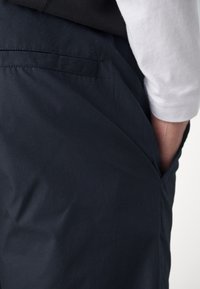 Main droite insérée dans la poche d'un pantalon bleu marine foncé, portant une chemise blanche à manches longues rentrée.