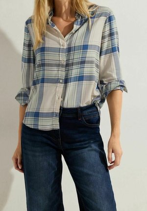 Femme portant une chemise à carreaux bleu clair et blanc à boutons avec manches retroussées et un jean bleu foncé, debout les mains détendues le long du corps.
