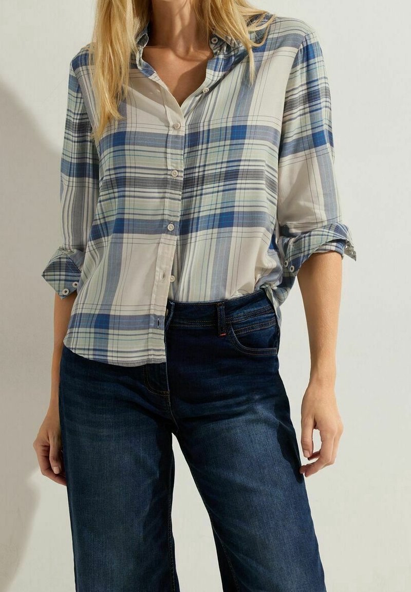 Femme portant une chemise à carreaux bleu clair et blanc à boutons avec manches retroussées et un jean bleu foncé, debout les mains détendues le long du corps.