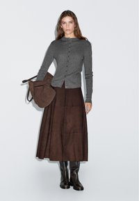 Gray ribbed cardigan with buttons, paired with a long brown suede skirt. Model holds a brown bag and wears knee-high black boots.